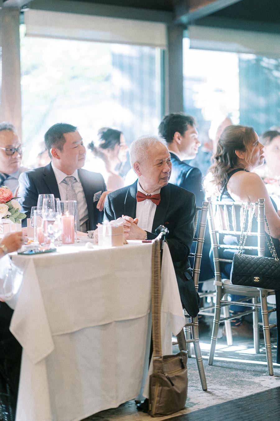 Wedding guests in formalwear sit at a bright reception table with candles, flowers, and silver chairs.