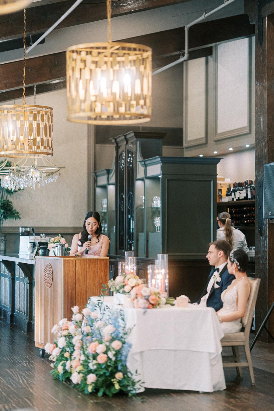 Woman gives a wedding reception speech at a podium while the bride and groom sit at the head table nearby.