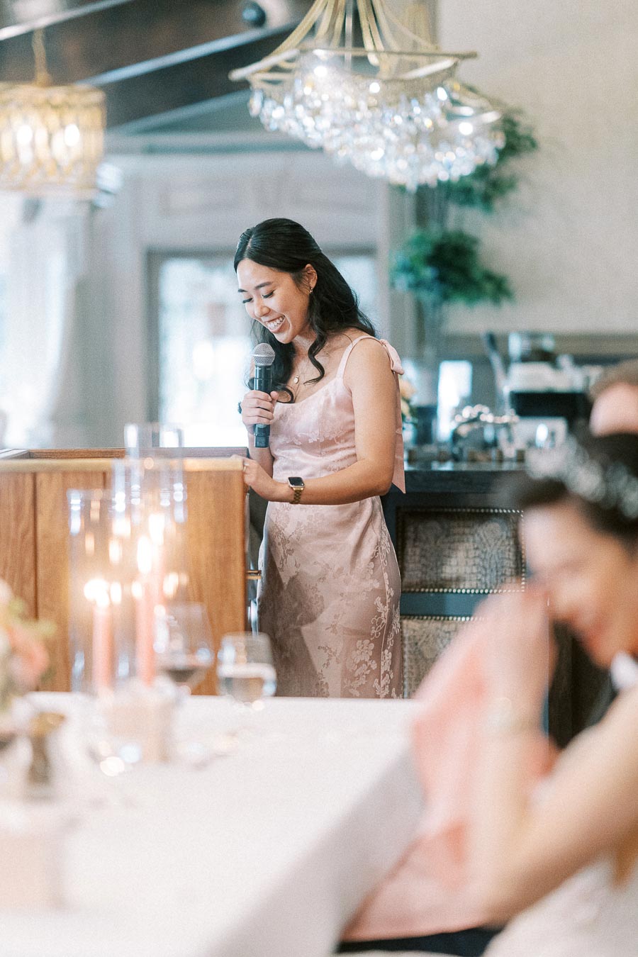 Woman in a pink dress smiles while giving a wedding reception speech with a microphone near chandeliers.
