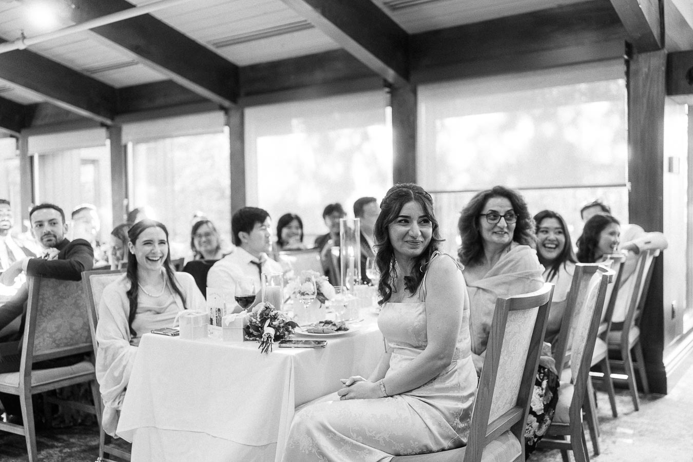 Wedding guests seated at a reception table smile and look across the room during speeches at Ancaster Mill.