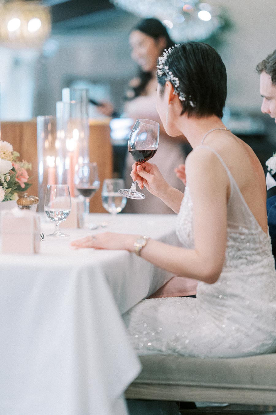 Bride in a beaded gown holds red wine at a candlelit reception table during a speech.