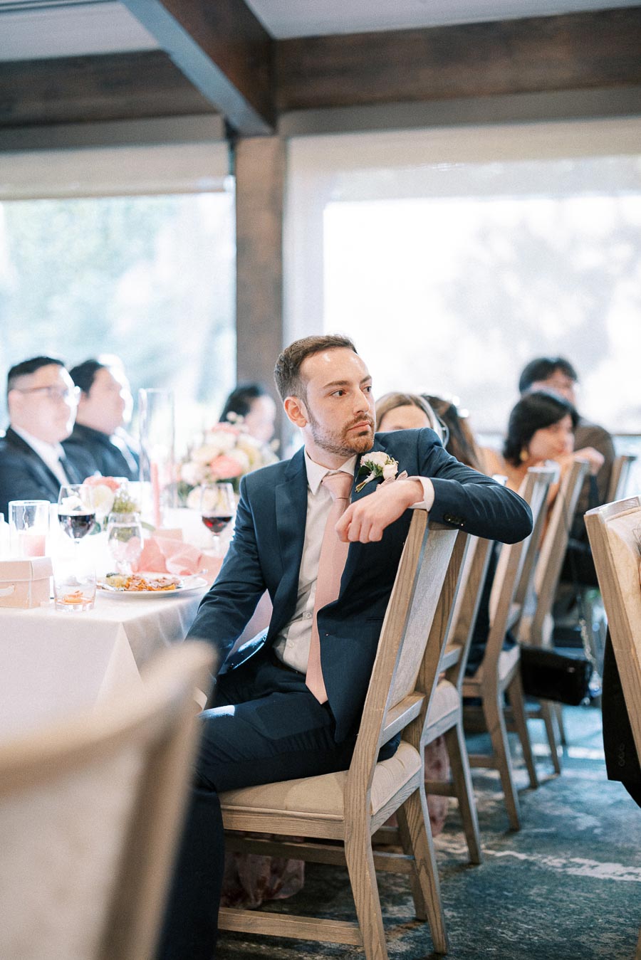 Wedding guest in a navy suit and pink tie seated at a reception table with guests and floral centrepieces