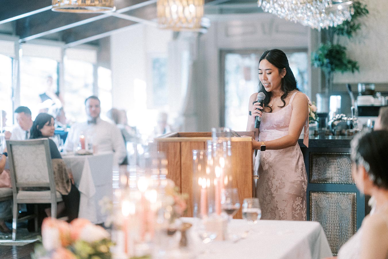 Woman in a pink dress gives a reception speech at a wooden podium as wedding guests listen in a bright dining room.
