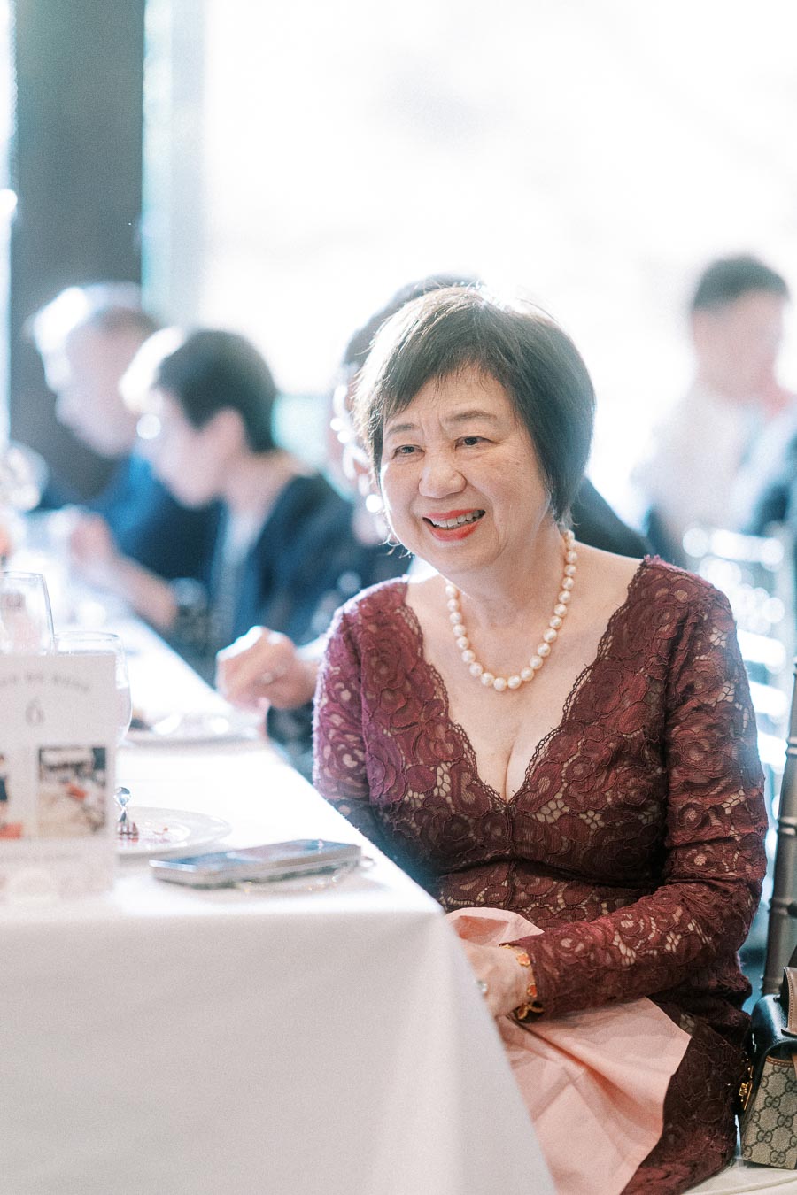 Smiling woman in a burgundy lace dress and pearl necklace seated at a wedding reception table