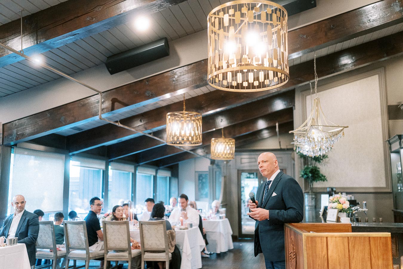 Man in a suit holds a microphone during a wedding reception speech in a dining room with chandeliers and seated guests.