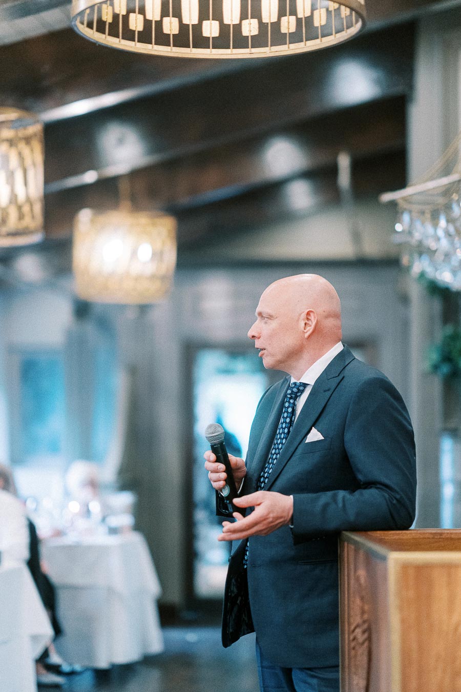 Man in a dark suit holds a microphone while speaking inside a chandelier-lit wedding reception room.