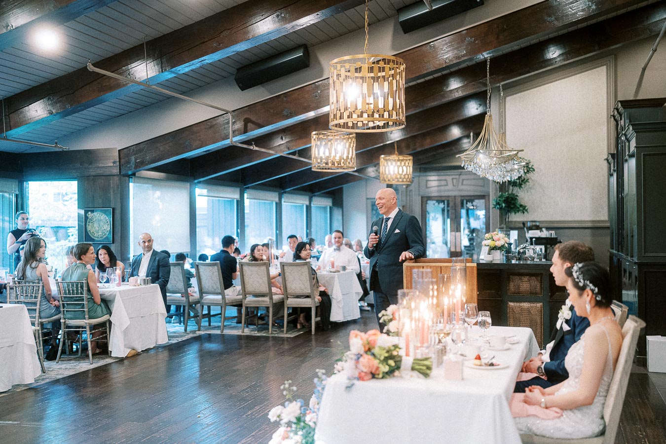 Man giving a wedding reception speech as Miffy and Rawle sit at the head table in a chandelier-lit dining room.