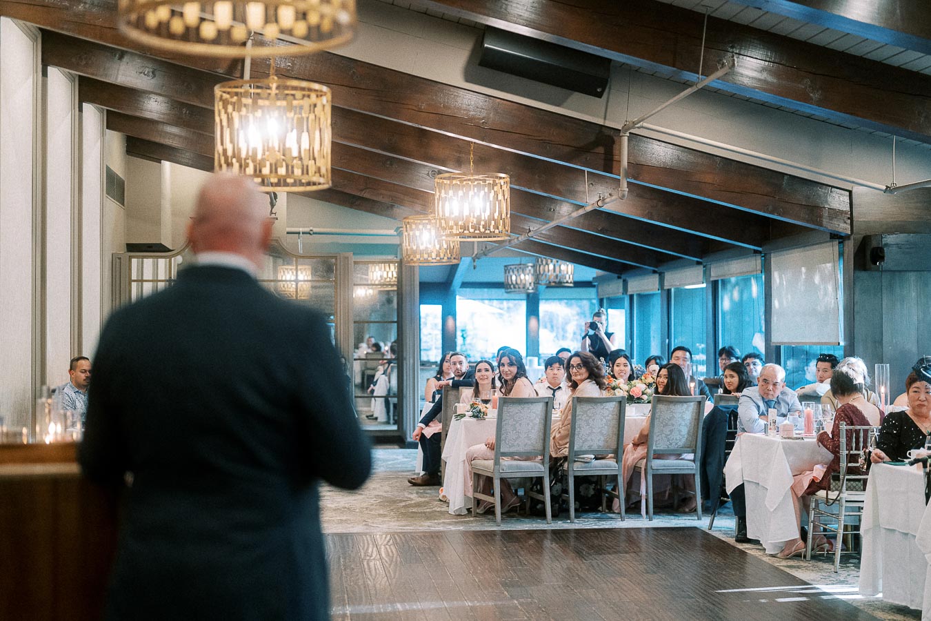 Guests seated at round reception tables watch a speaker in a warmly lit room with chandeliers and wood beams.