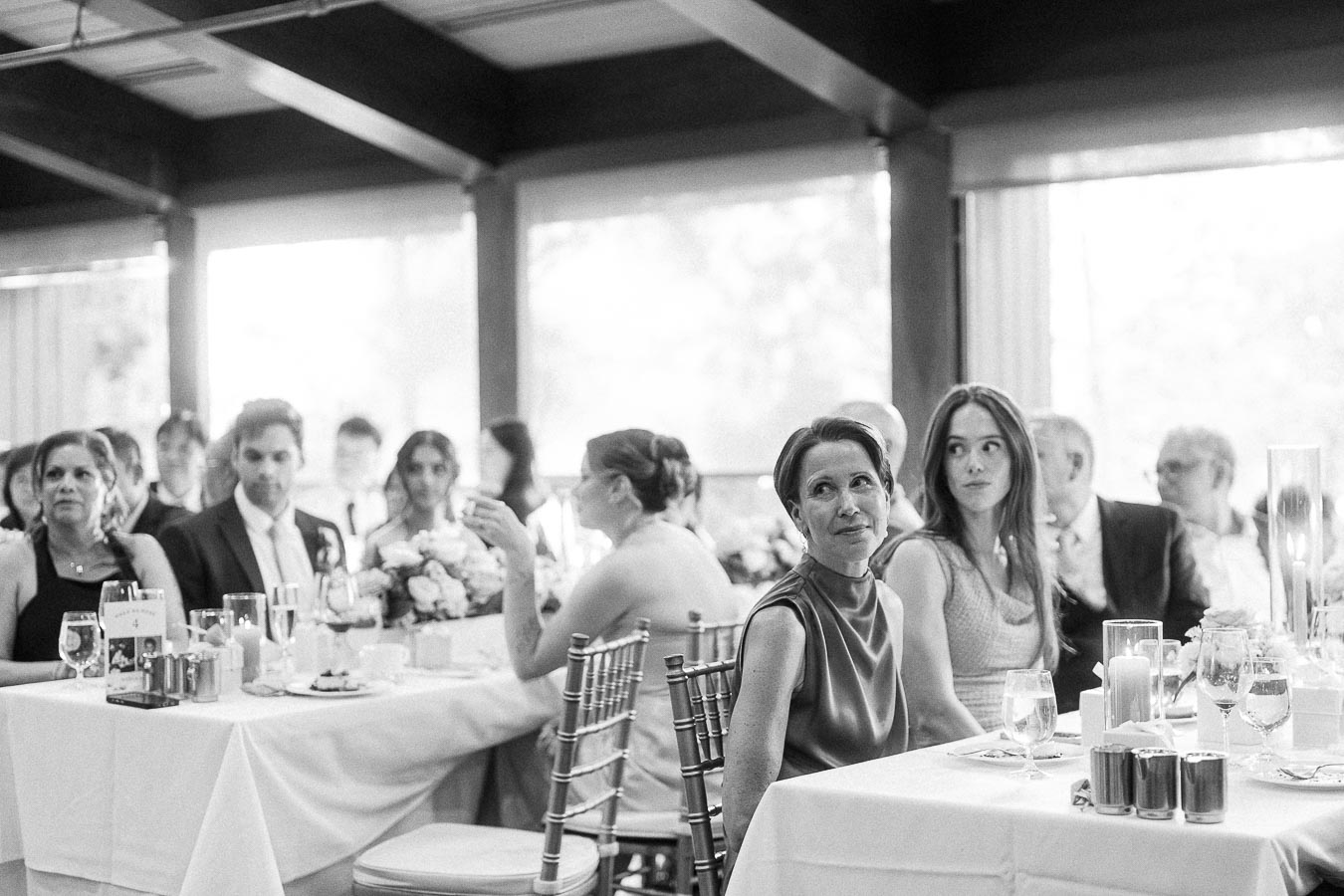 Black-and-white view of wedding reception guests seated at tables with candles, drinks, and floral centerpieces.