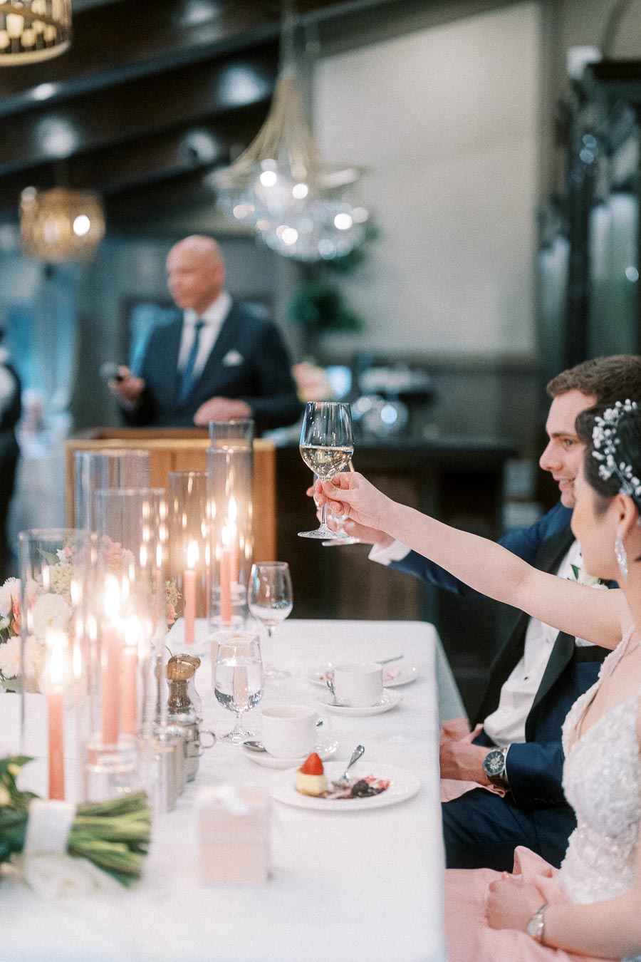 Bride raises a wine glass beside the groom at a candlelit head table during a wedding speech.