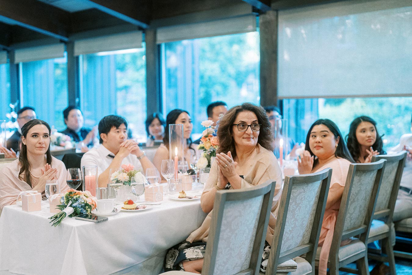 Wedding reception guests seated at tables clap beside candles, flowers, wine glasses, and large windows.