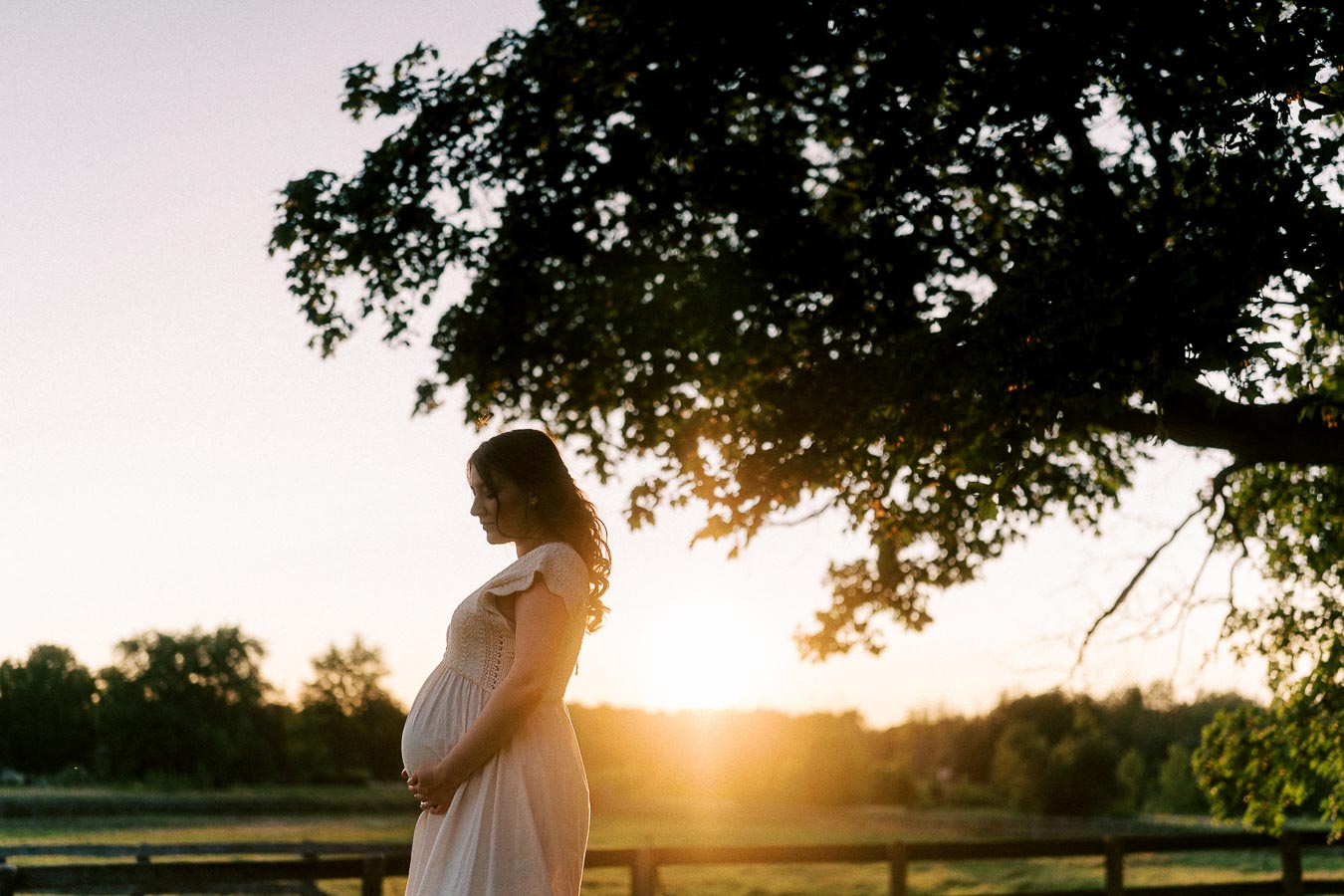Pregnant woman in a white dress standing beneath a tree at sunset