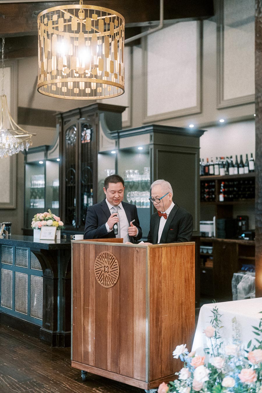 Two men stand at a wooden lectern with a microphone during a wedding reception speech in a chandelier-lit room.