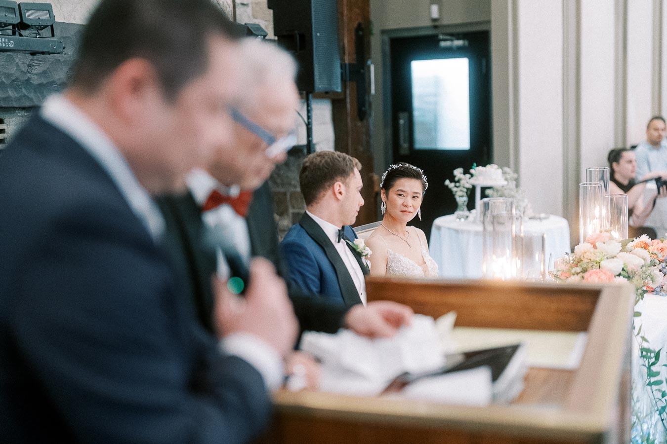 Miffy and Rawle seated at their reception table listening to a speech in a softly lit dining room