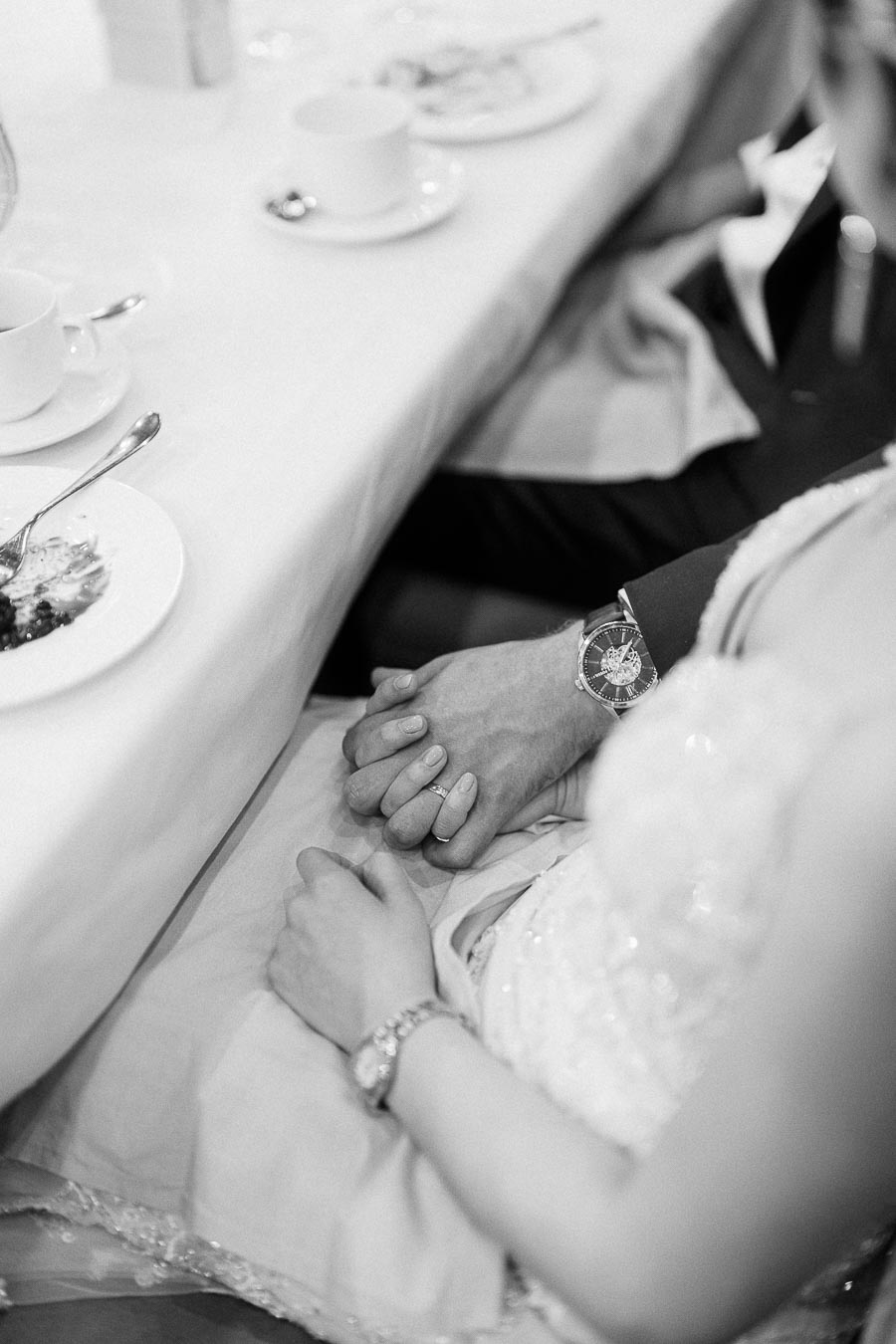 Black and white close-up of Miffy and Rawle holding hands beneath a reception table with tea cups and dessert plates.