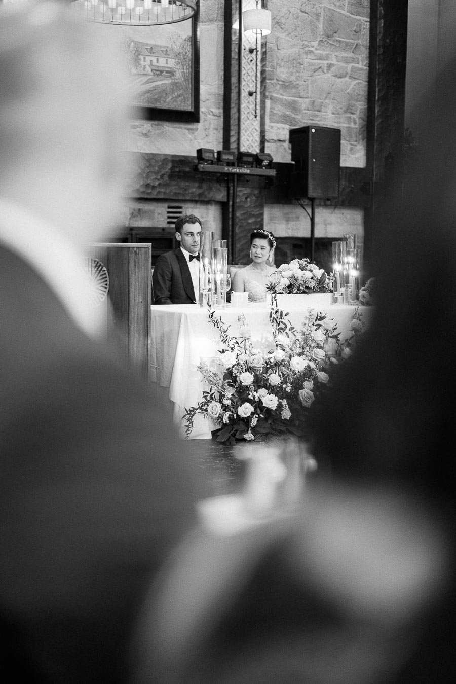 Miffy and Rawle seated at their reception head table with candles and flowers inside a stone-walled room.