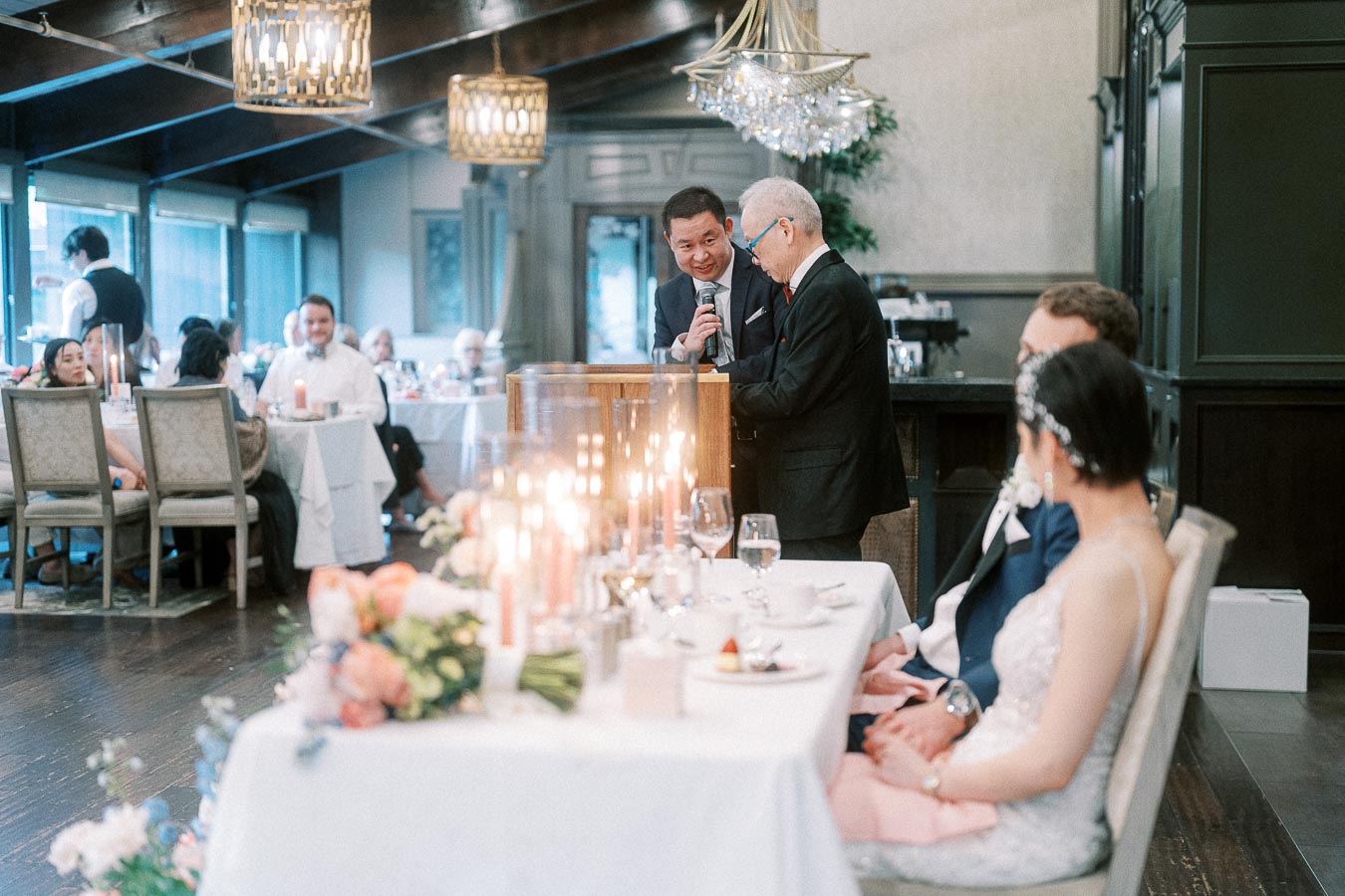 Two men give a reception speech while the couple sit at the head table with candles and flowers.