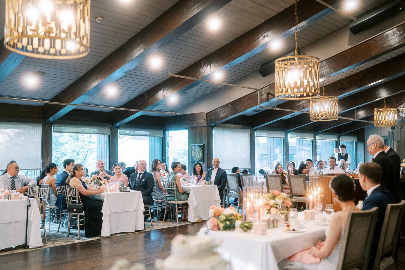 Wedding guests seated at reception tables listen to a speech in a bright dining room with wooden beams and chandeliers.