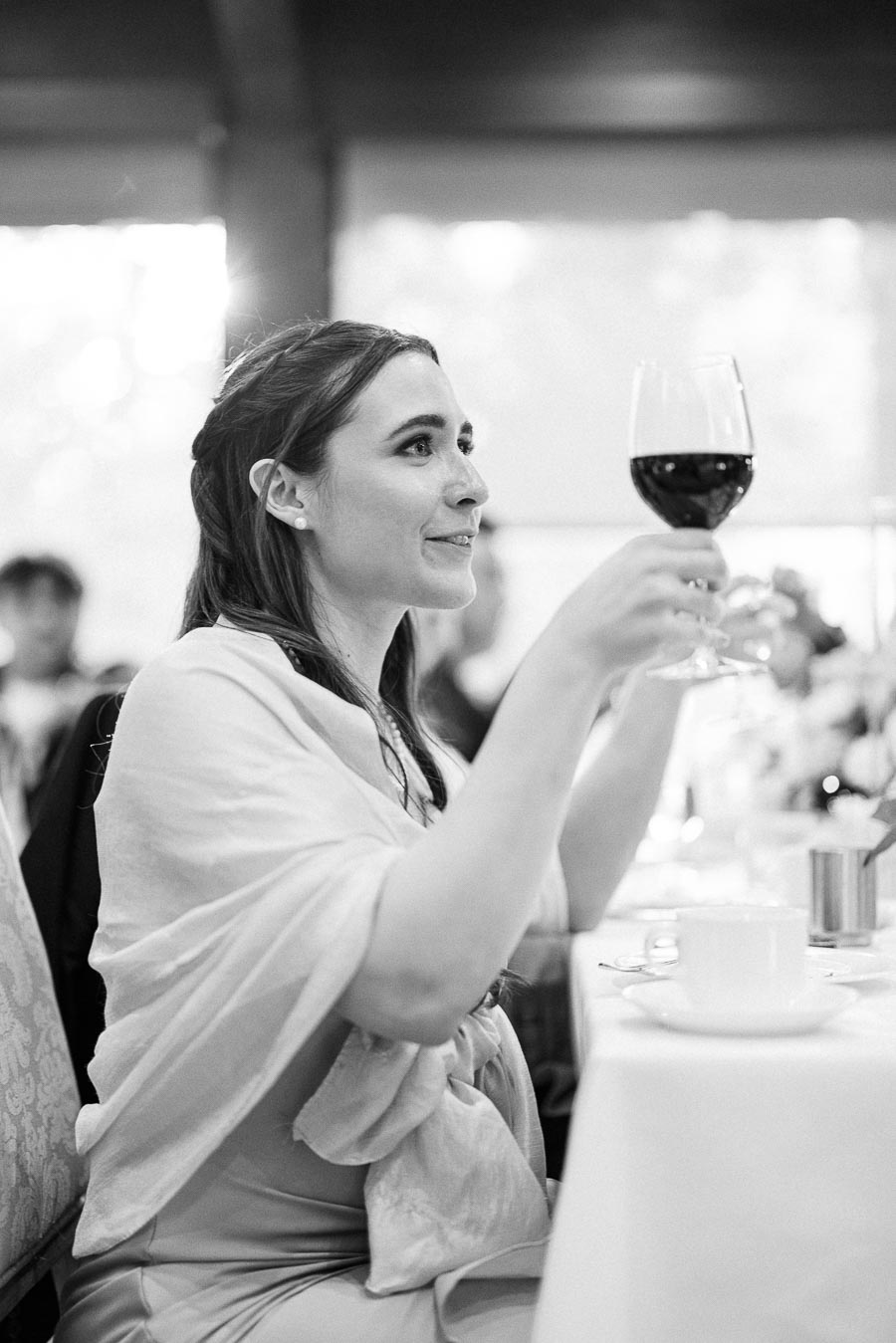 Guest seated at a reception table raises a glass of red wine during a wedding toast.