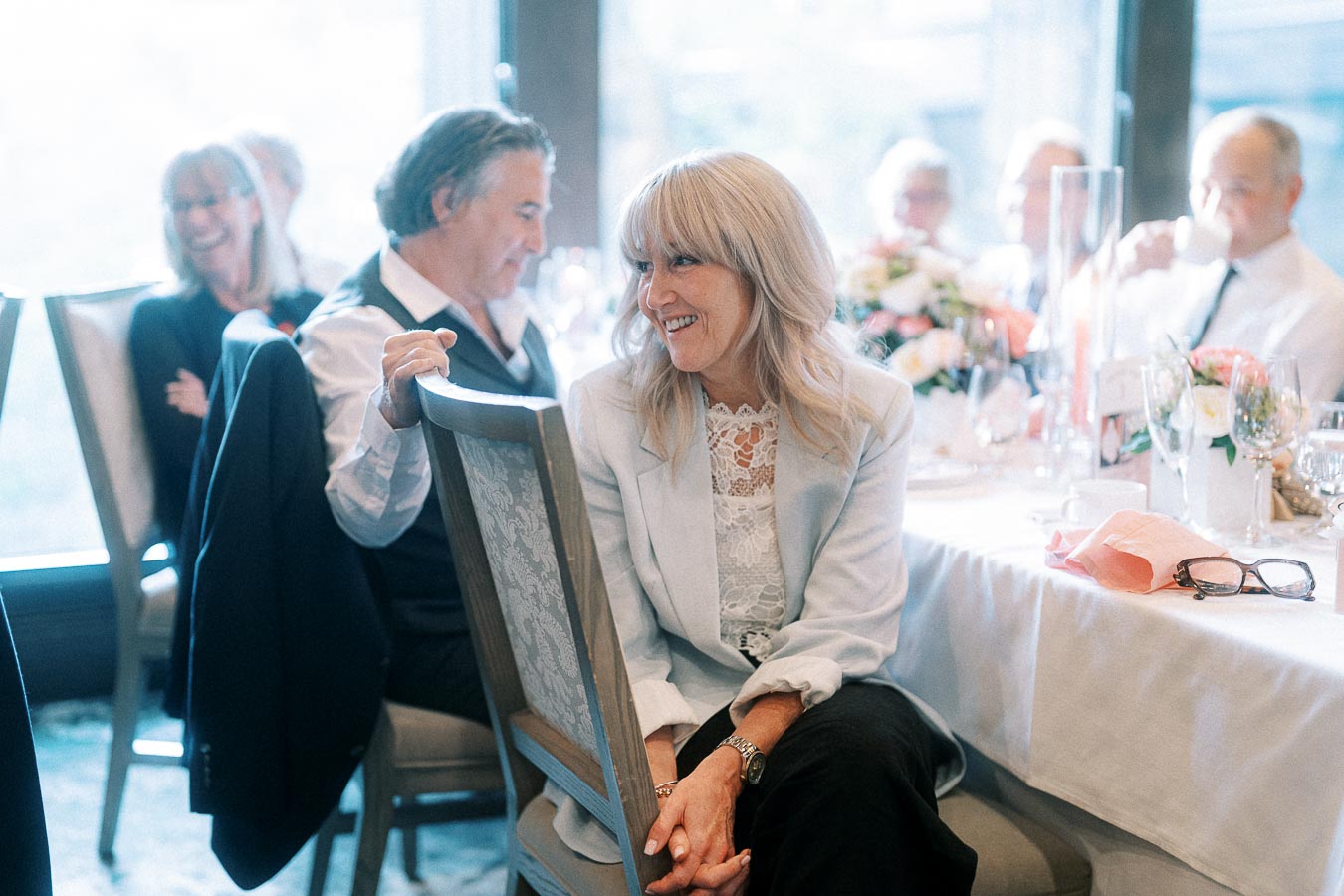 Smiling guest seated beside a reception table with flowers, glassware, and other guests in soft window light.