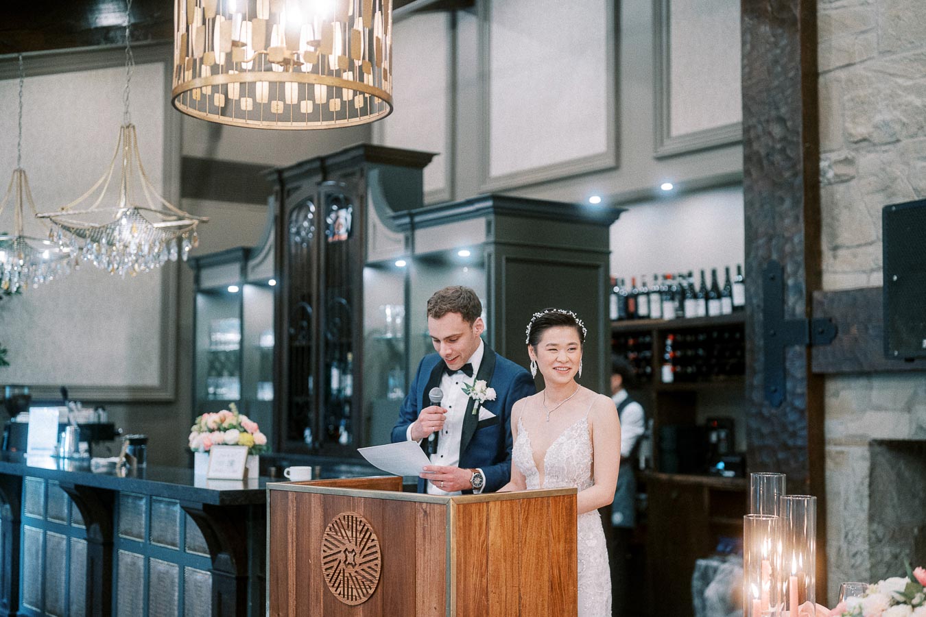 Rawle reads from a paper at a reception podium while Miffy smiles beside him in her wedding dress.