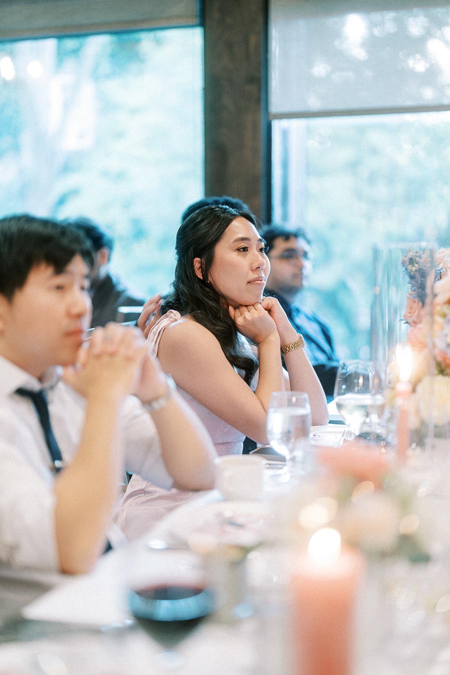 Woman in a pale dress sits thoughtfully at a candlelit wedding reception table with guests nearby.