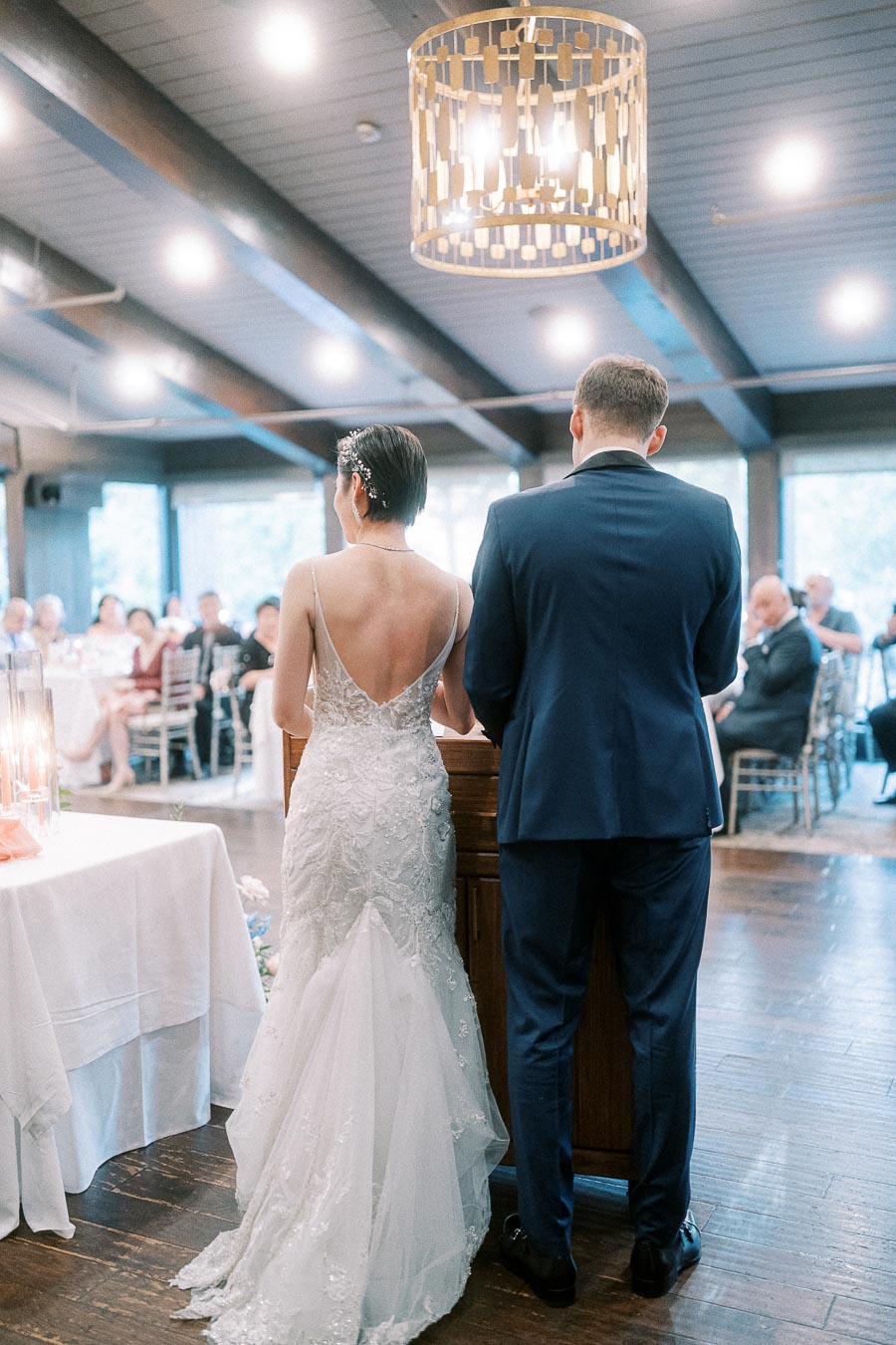Bride and groom stand at the ceremony lectern facing guests in a bright timber-ceiling room.