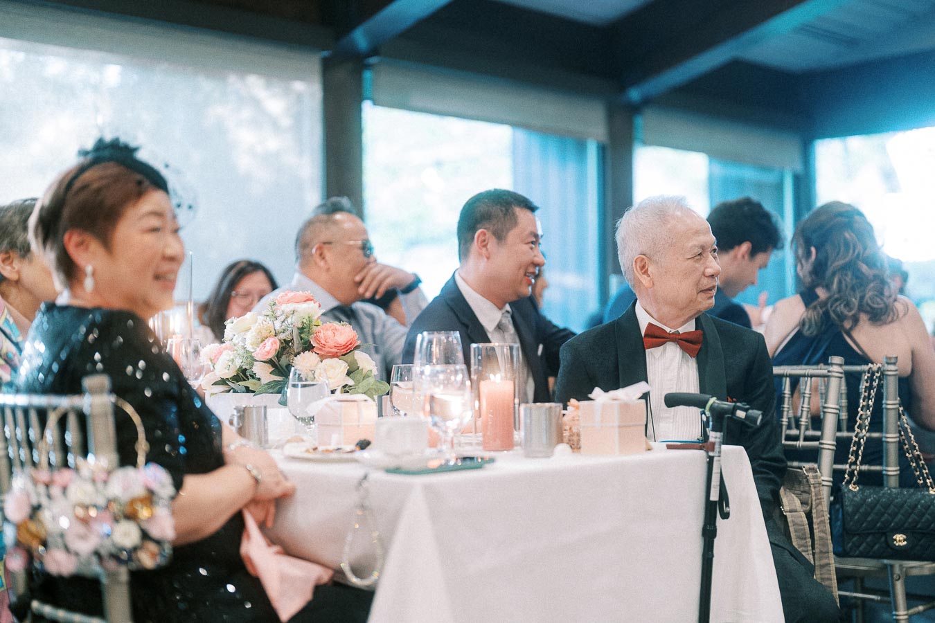 Wedding guests seated at a reception table with candles, flowers, glassware, and an older man in a red bow tie.