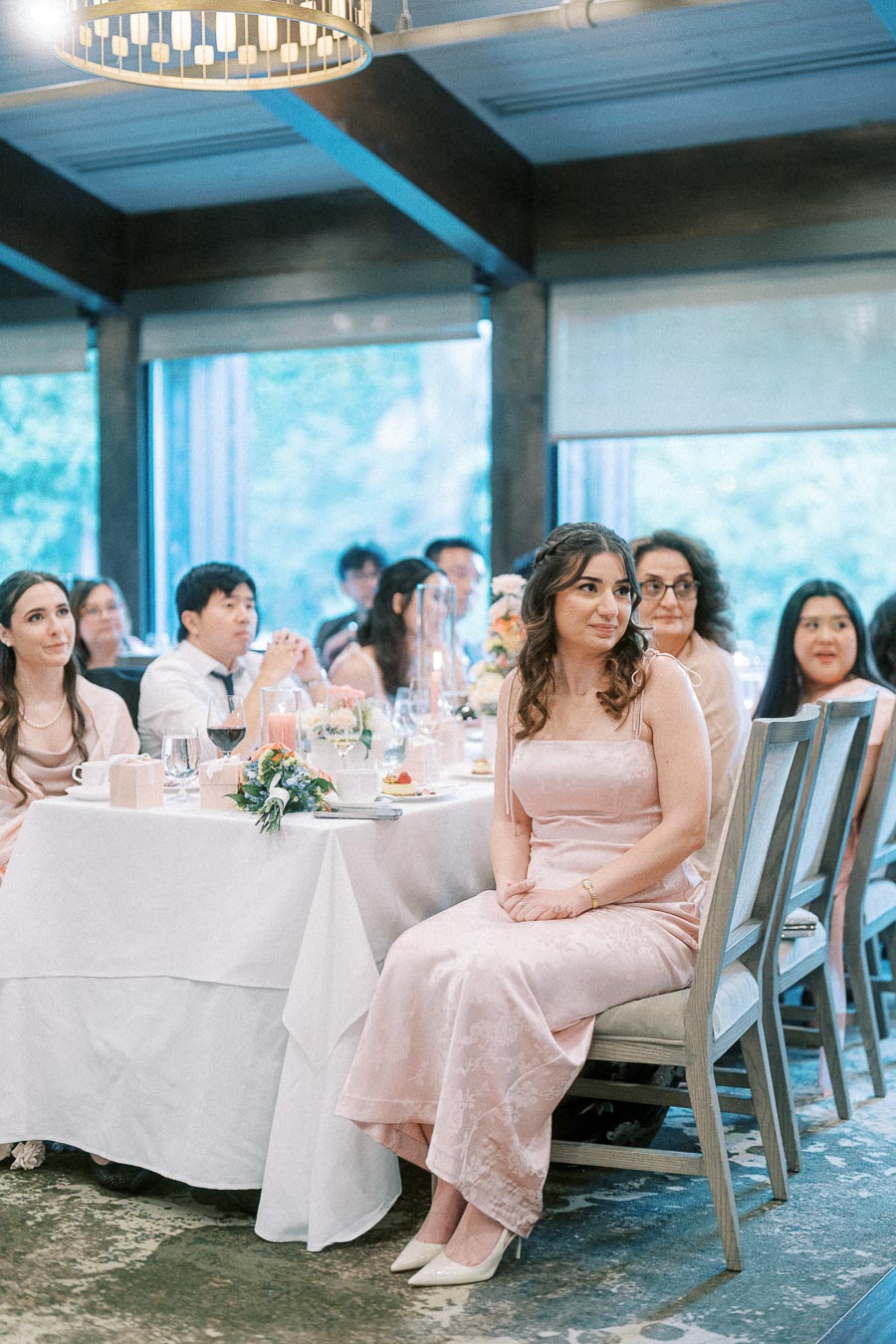 Wedding guests seated at a reception table, with a woman in a pale pink dress turned toward the room.