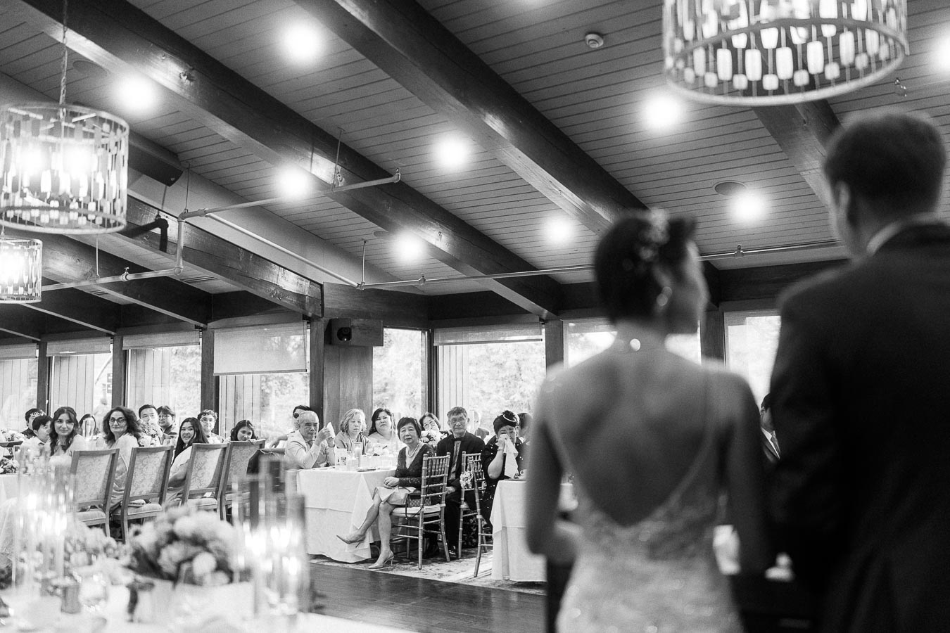 Bride and groom stand before seated reception guests in a timber-beamed dining room at Ancaster Mill.