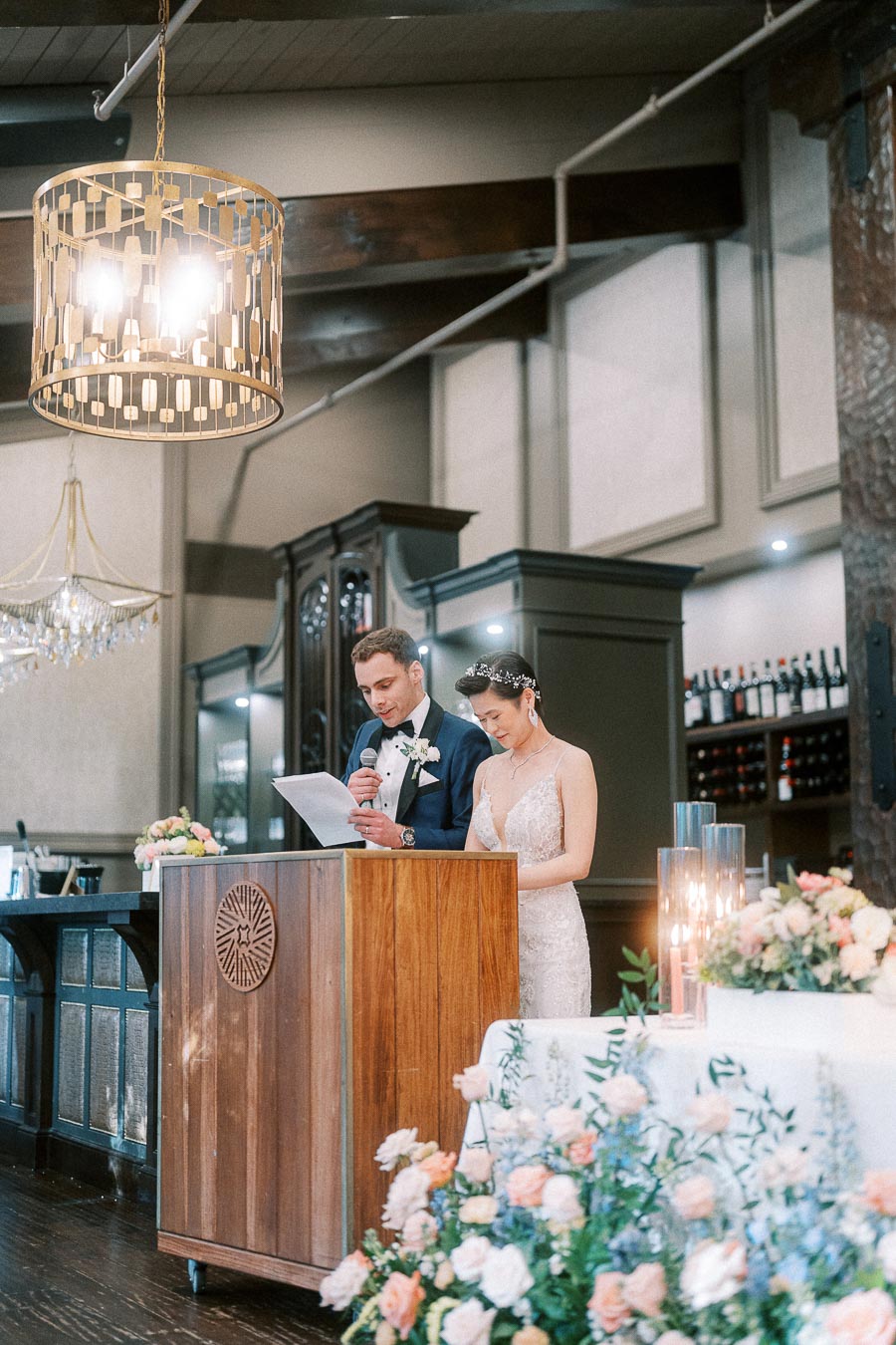Rawle reads from a paper at a wooden podium while Miffy stands beside him during their wedding reception.