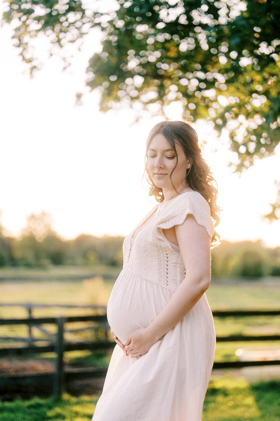 Pregnant woman in a cream dress cradles her belly by a farm fence