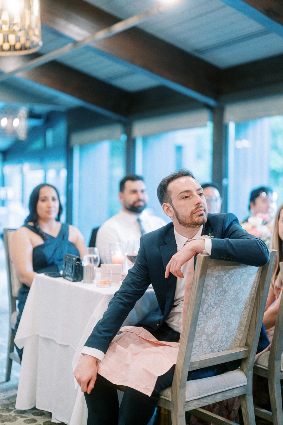 Man in a dark suit sits turned in a reception chair while guests listen at nearby tables.