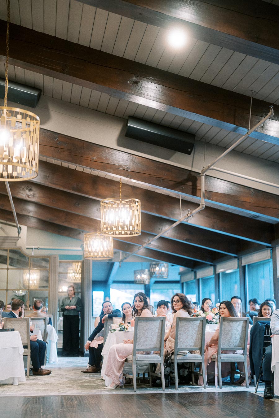 Wedding guests seated at reception tables under wood beams and glowing chandeliers at Ancaster Mill