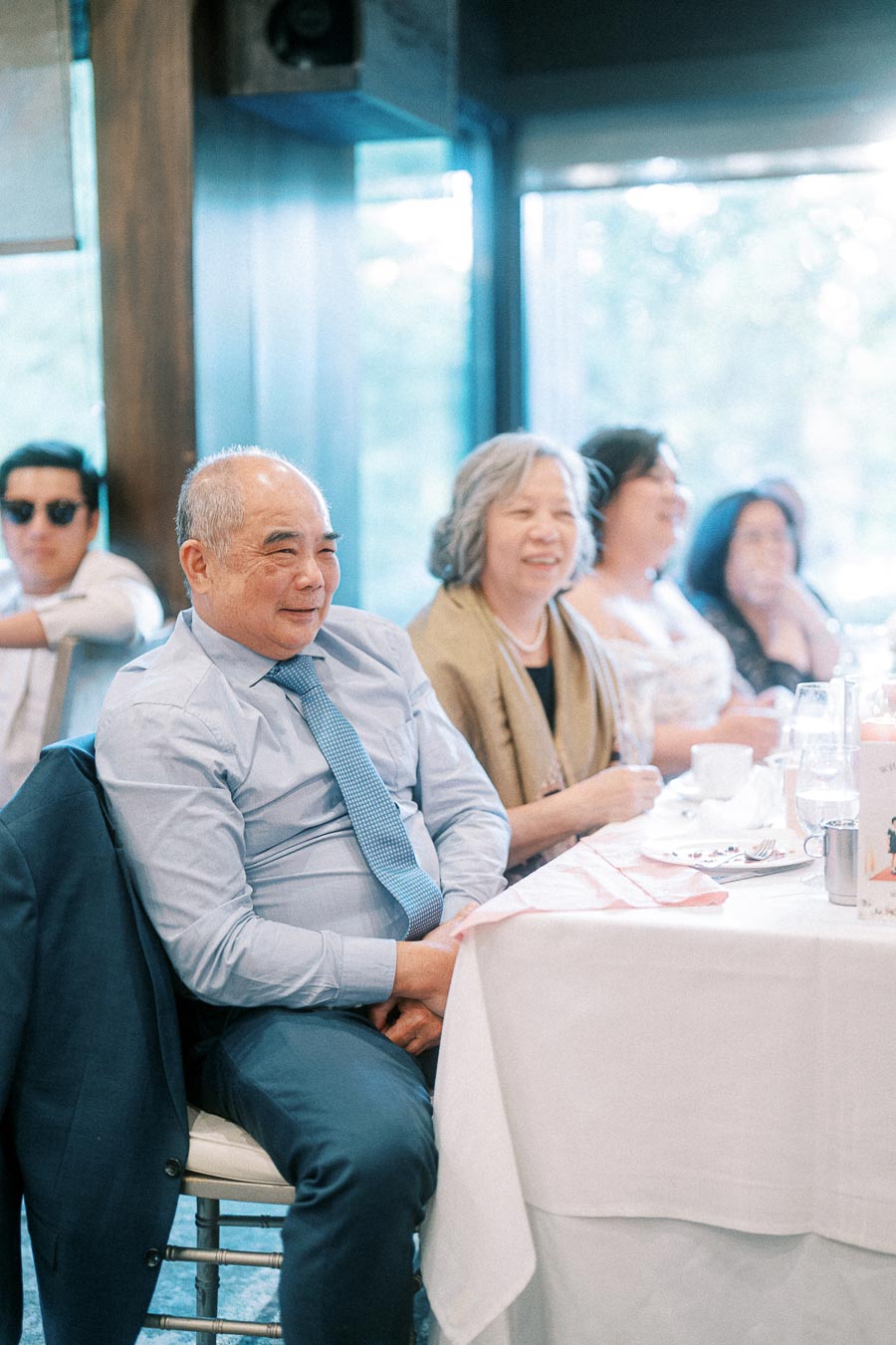 Wedding guests seated at a reception table, smiling and listening near bright windows.