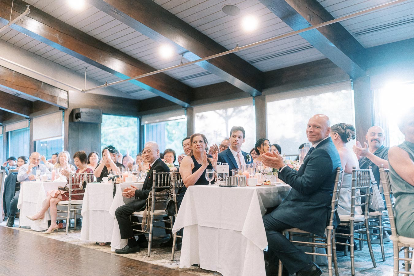 Wedding reception guests seated at round tables applaud inside a bright beamed dining room at Ancaster Mill.
