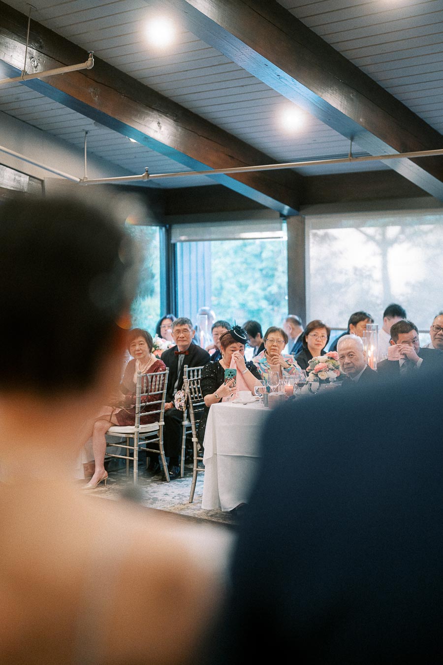 Wedding guests seated at round tables watching speeches in a reception room with wood beams and large windows.