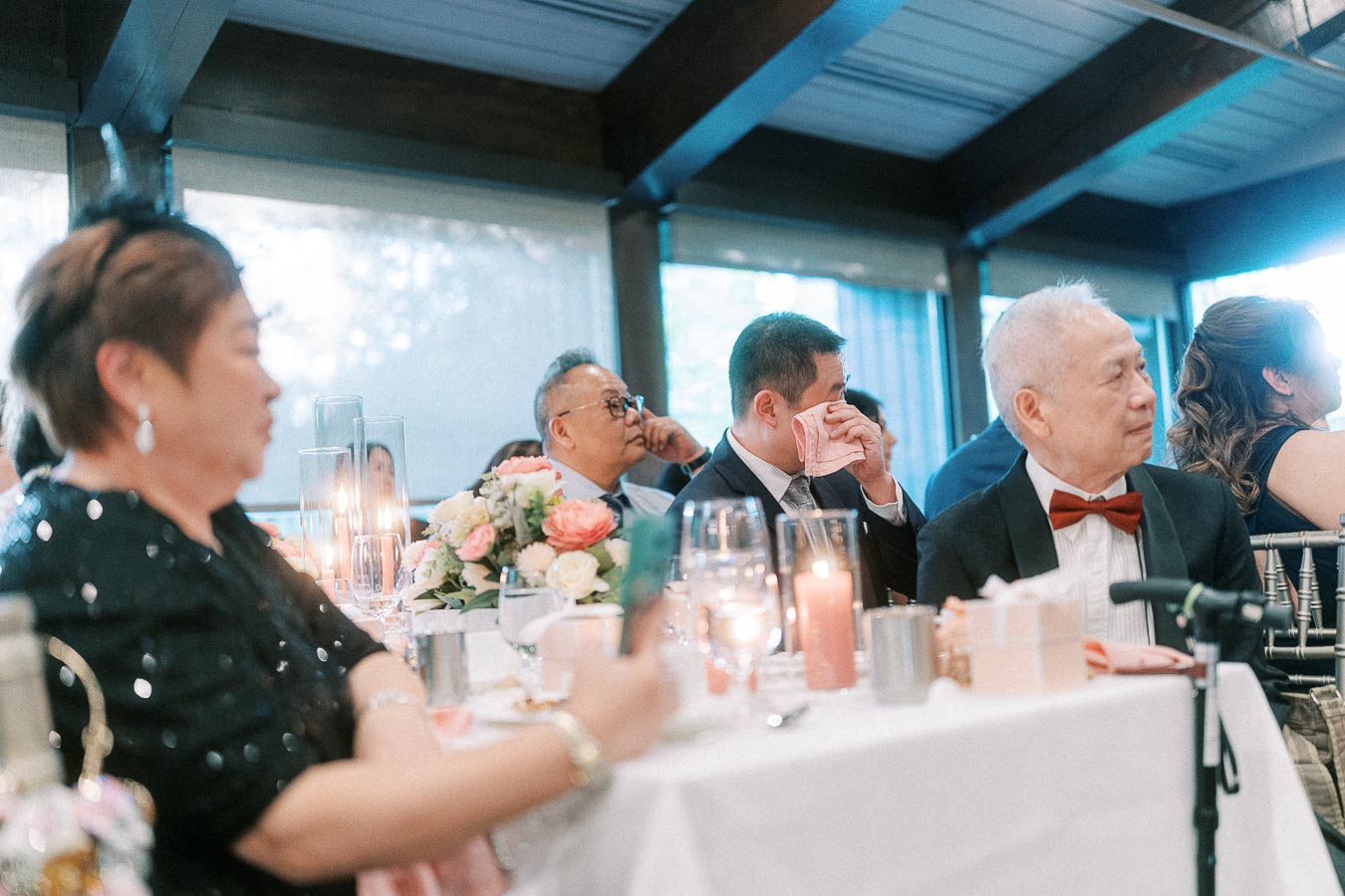 Wedding guests seated at a candlelit reception table, with one man wiping his eyes during speeches.