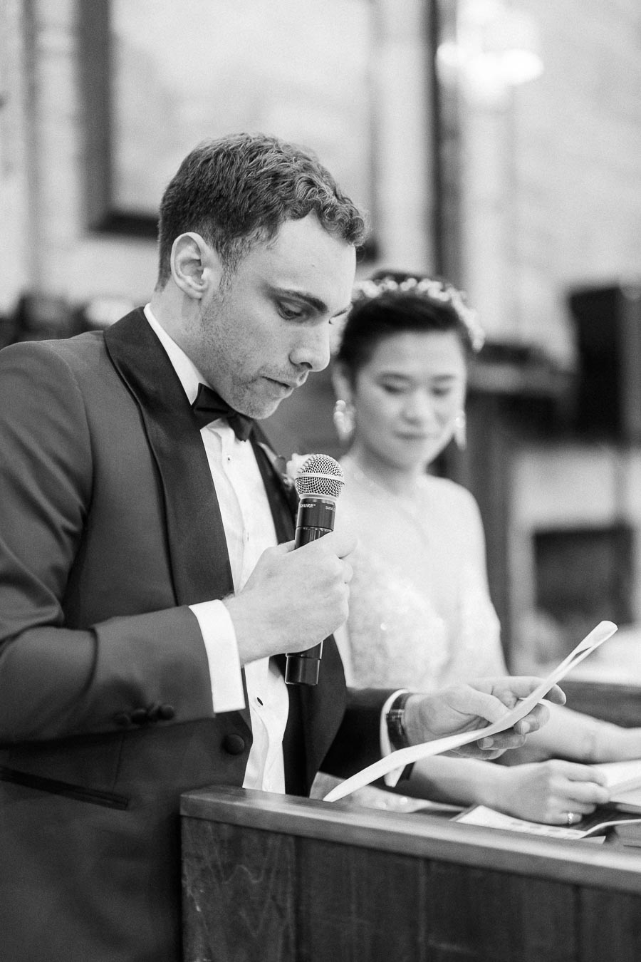 Groom reads from a paper into a microphone while bride stands beside him during their wedding ceremony.