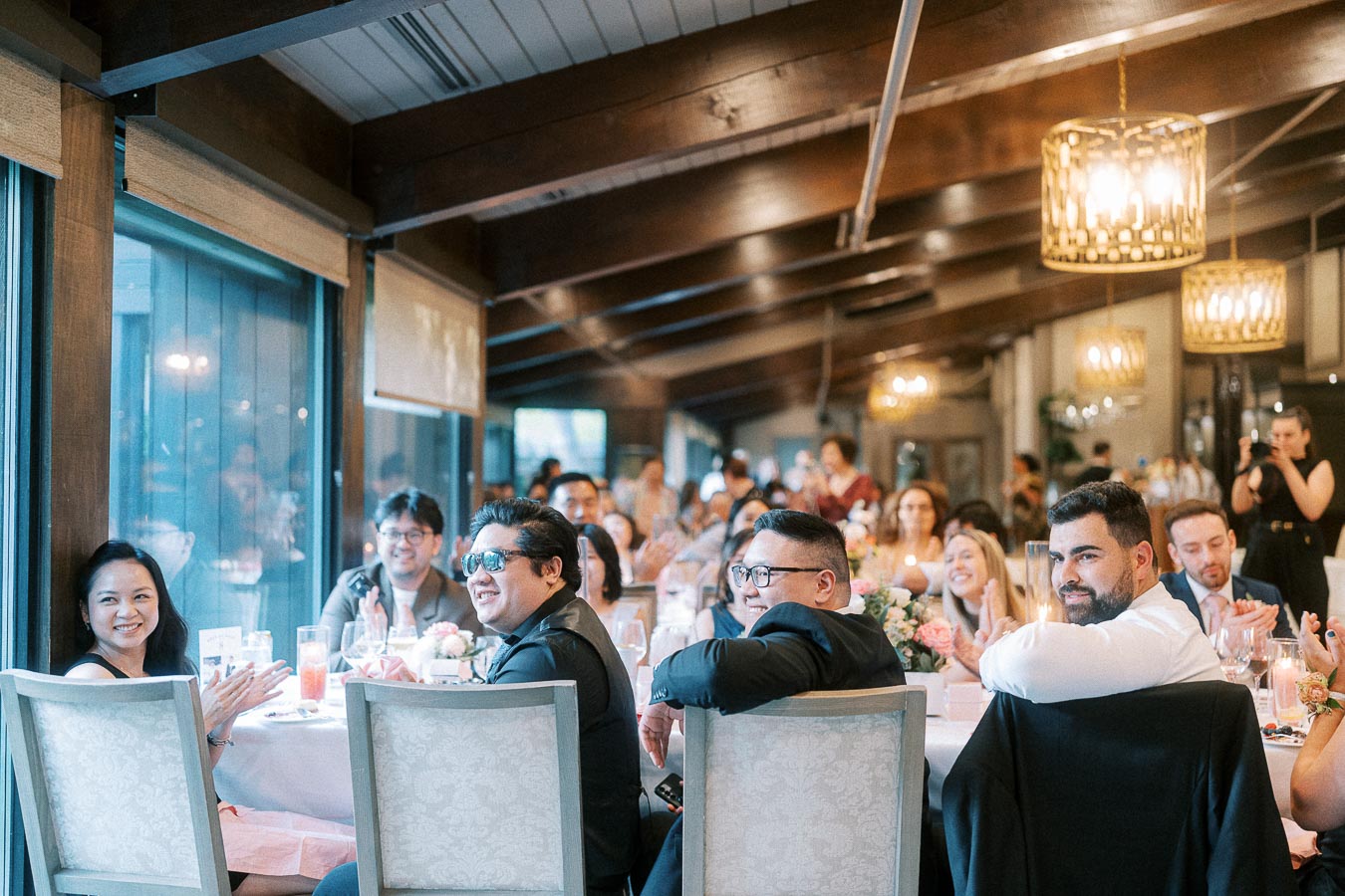Wedding reception guests seated at long tables look toward the camera in a warmly lit banquet room.