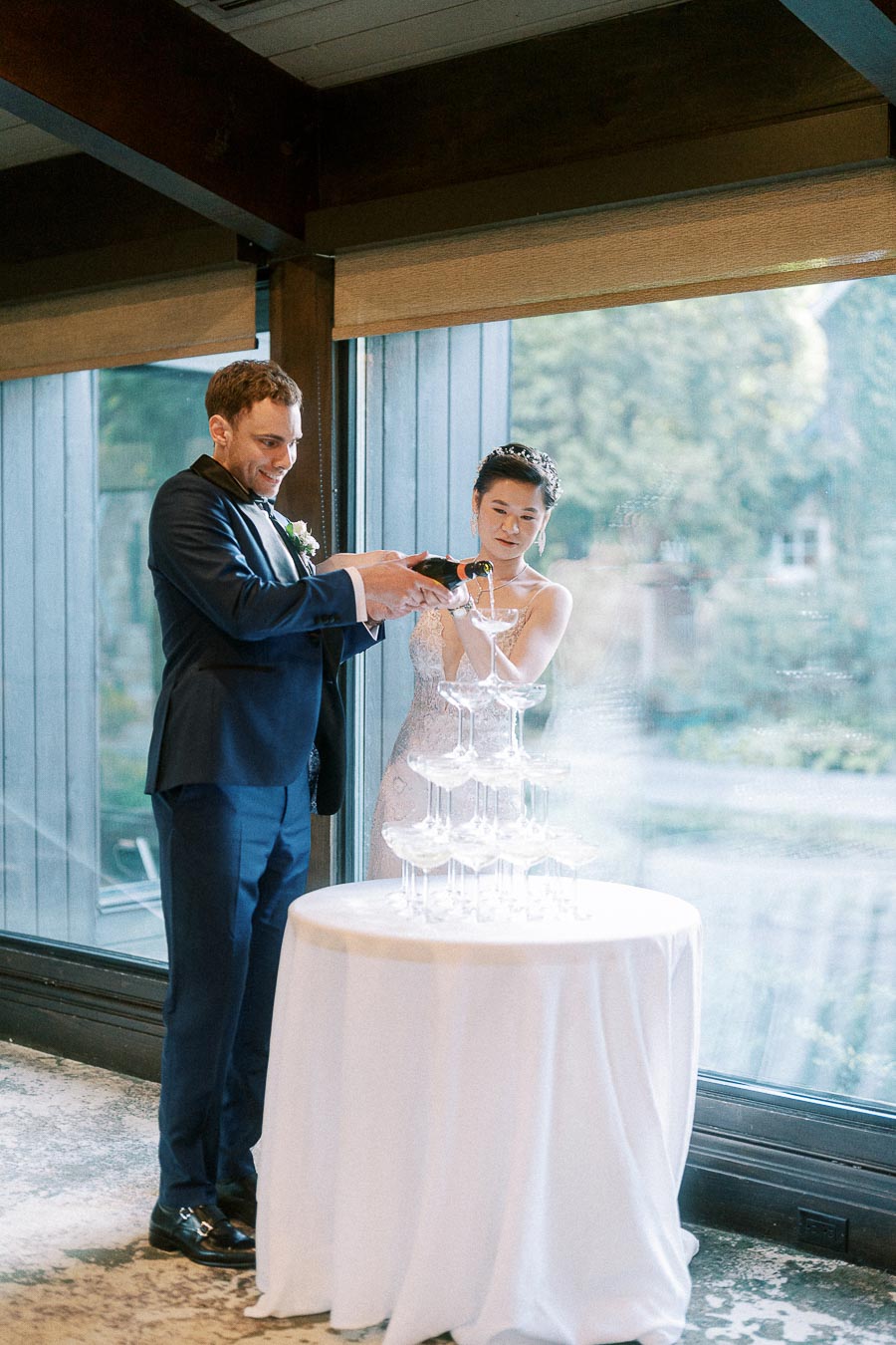 Miffy and Rawle pour champagne over a coupe glass tower beside large windows at their reception.