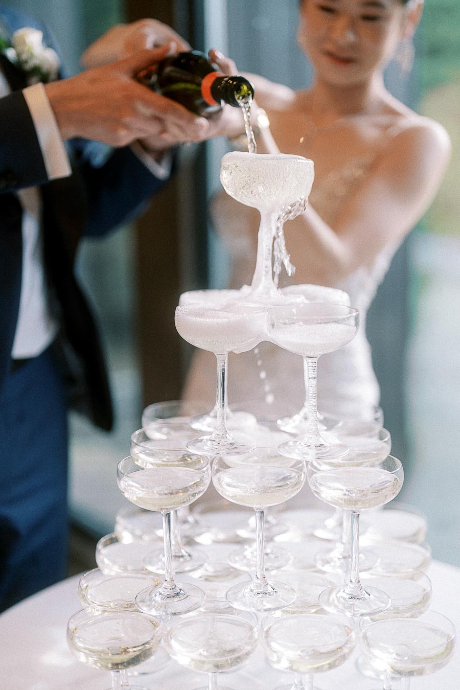 Bride and groom pour champagne into a coupe tower, with bubbles spilling over the top glass.