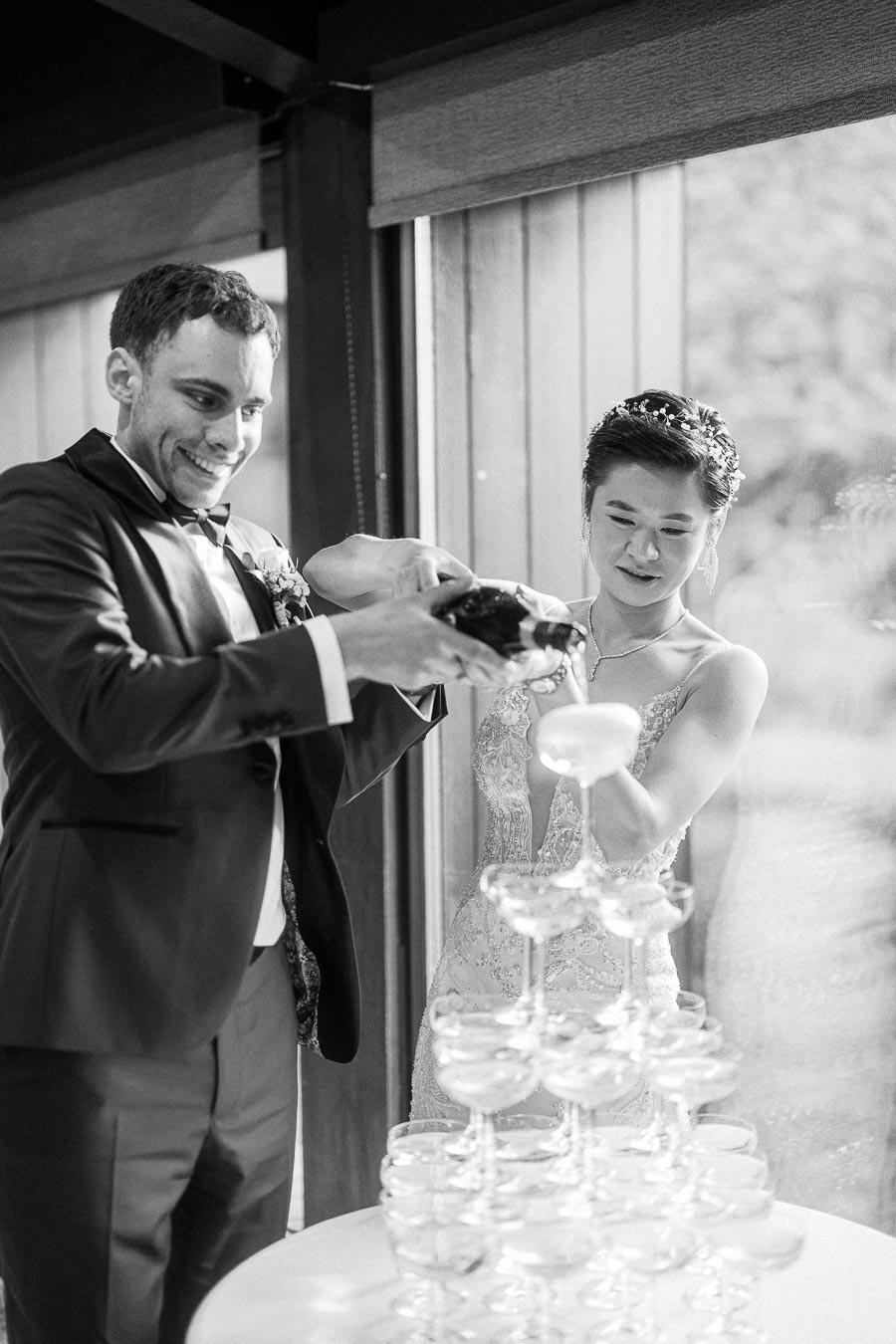 Miffy and Rawle pour champagne into a coupe tower beside a window during their wedding reception.