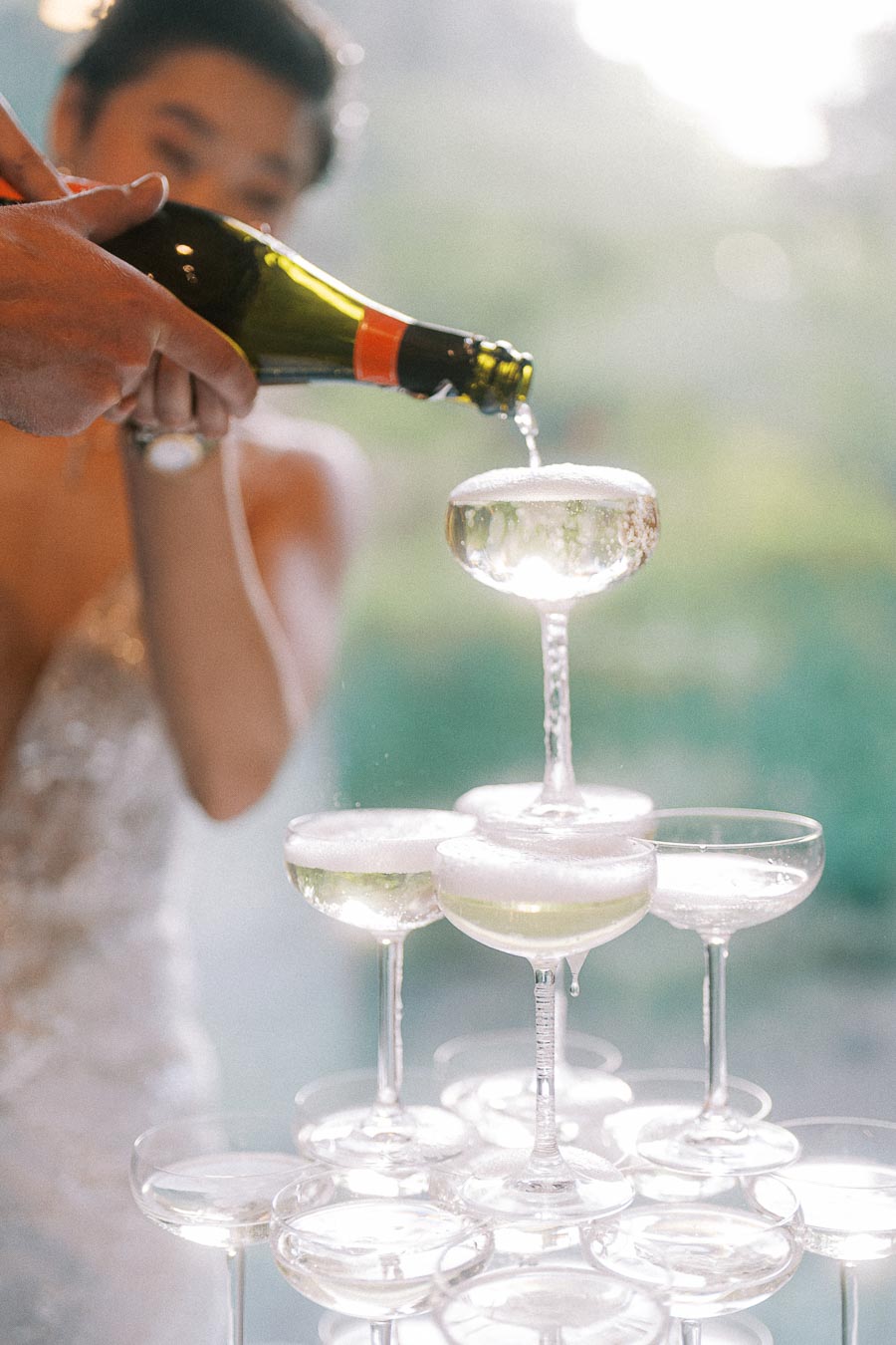 Bride pours champagne into a coupe glass at the top of a sparkling tower in soft afternoon light.