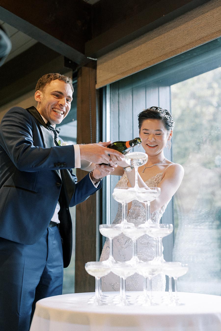 Couple joyfully pouring champagne into a cascading tower of glasses at their wedding reception, showcasing a joyful celebration.