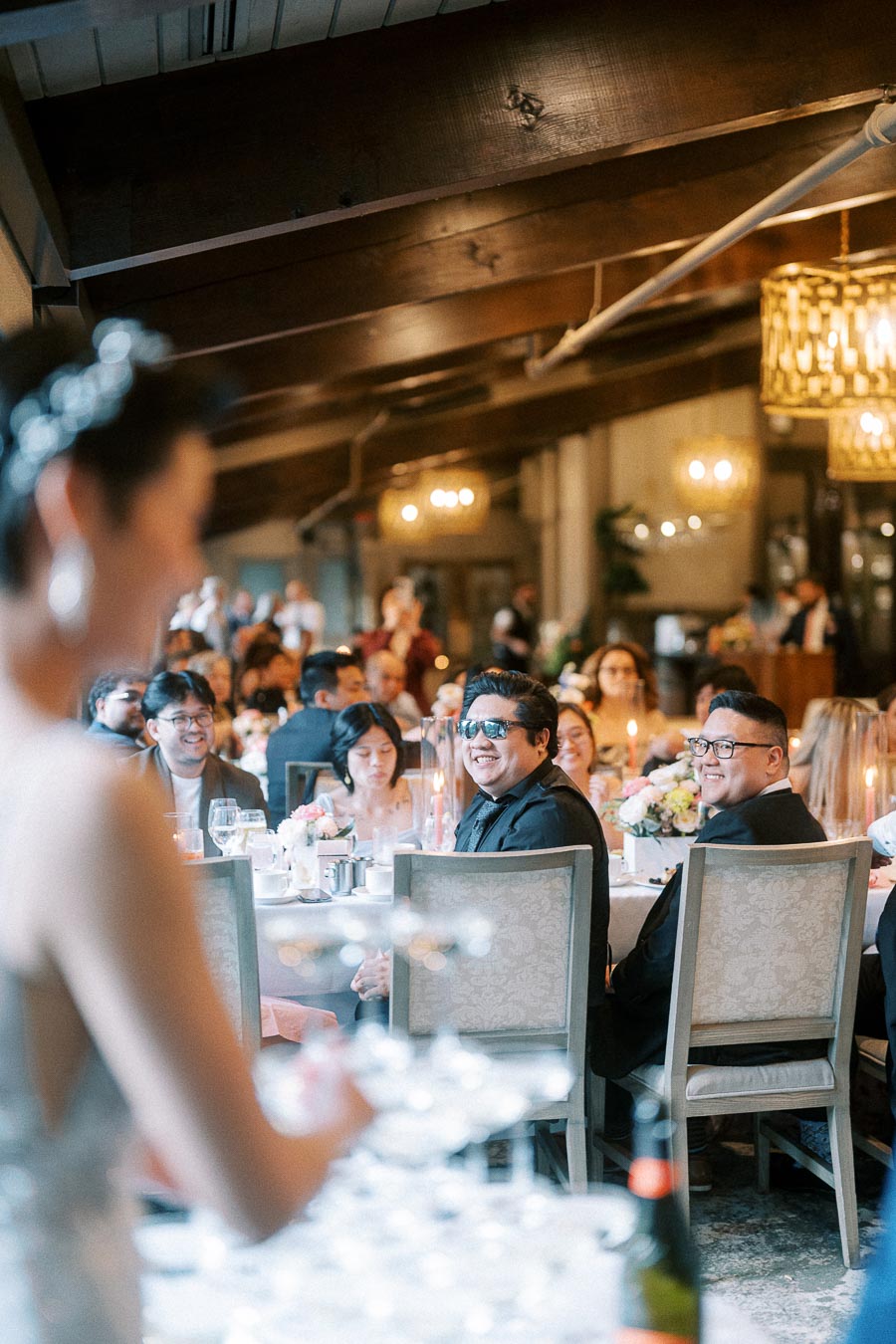 Elegant wedding reception with guests seated at tables adorned with floral arrangements and a champagne tower, under warm, decorative lighting.