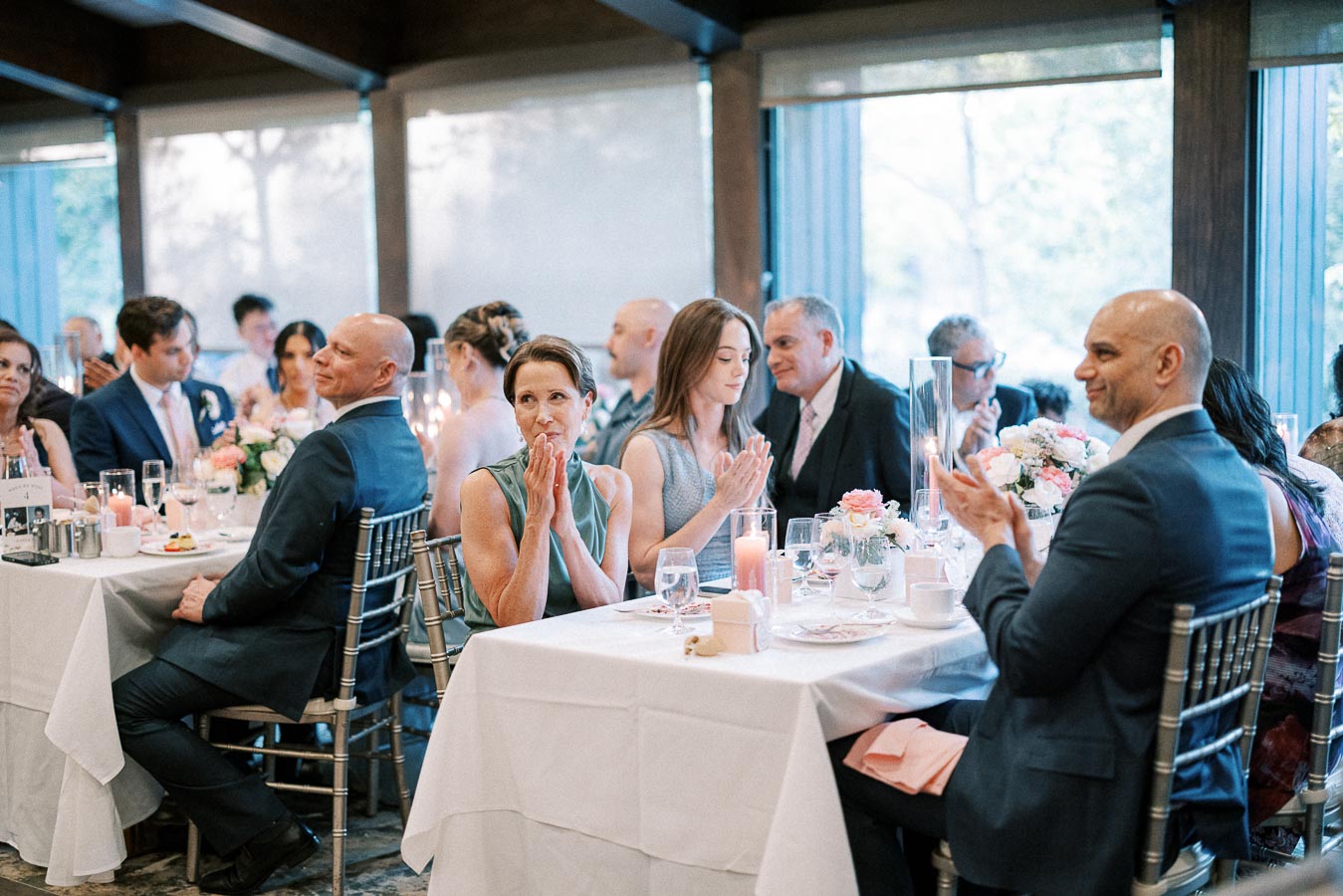 A group of elegantly dressed guests seated at beautifully decorated tables, clapping during a formal event or wedding reception, surrounded by floral centerpieces and ambient lighting.