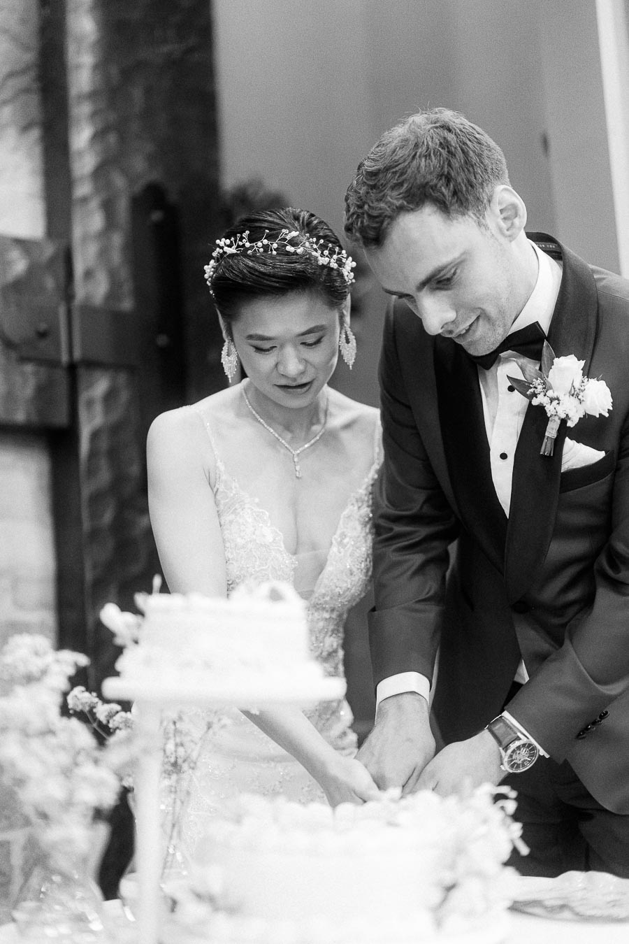 A bride and groom elegantly cutting their wedding cake together in a black and white photograph, highlighting their formal attire and the celebratory ambiance.