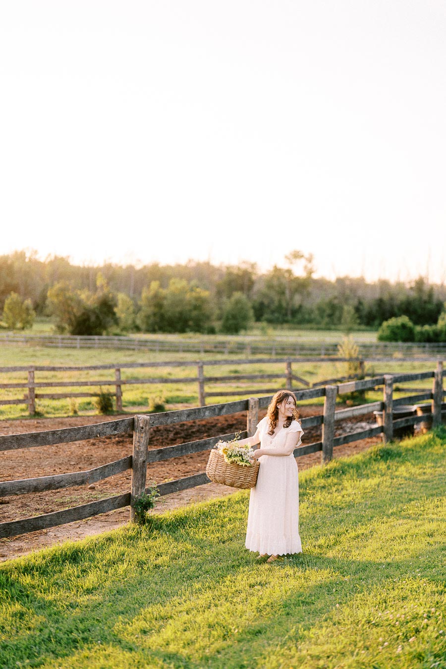 Pregnant woman in a white dress holding a flower basket beside farm fencing