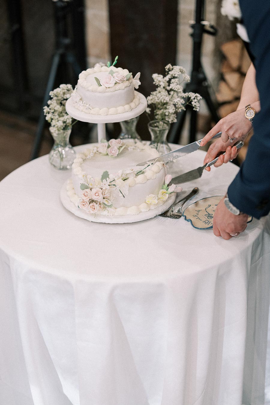 Two-tiered white wedding cake with floral decorations being cut by a person at a reception, surrounded by delicate vases of flowers on a white tablecloth.